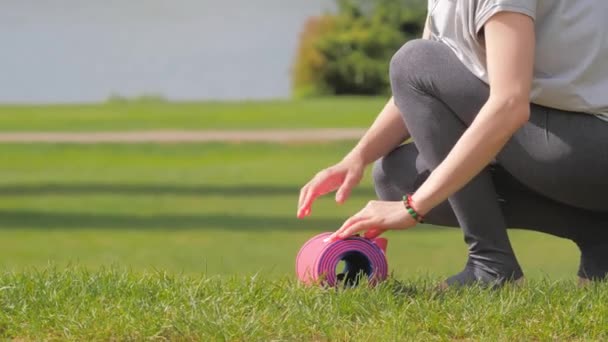 Обрезанный вид молодой женщины в спортивной одежде распространяется фитнес-мат на зеленой траве — стоковое видео