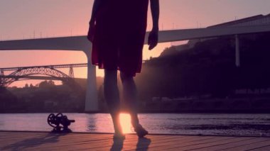 İskelede kırmızı elbiseli genç bir kadın gün batımında Porto Köprüsü manzarasına karşı dans ediyor.