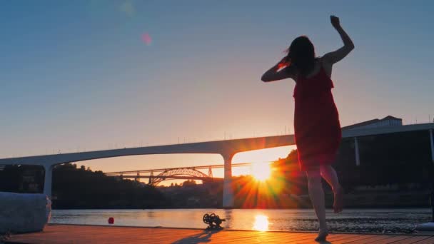Женщина в красном платье на пирсе танцует и веселится перед видом на городские мосты — стоковое видео
