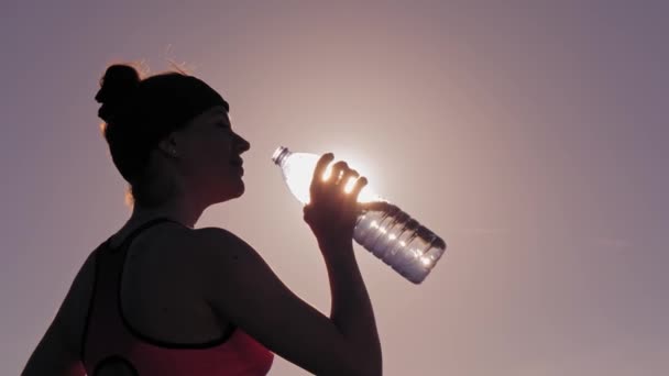 Attractive sportwoman drinks water from a bottle against sunlight — Stock Video