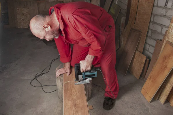 车间里的木匠用锯子锯断层压板 穿着红色工作服 配有电动工具的工人会割断花束 工人用电锯切割木板 灰尘四处飞扬 — 图库照片