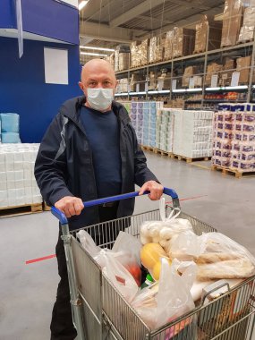 Koruyucu maskeli, alışveriş arabalı, süpermarkette ürünleri olan bir adam. Coronavirus covid enfeksiyonu 19 salgını sırasında süpermarkette tramvayla maskeli bir adam. Bir salgında sosyal uzaklık ve alışveriş