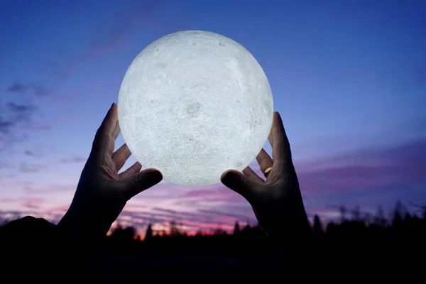 Round full moon in hands against evening sky. Lunar model, moon-shaped lamp with moon craters