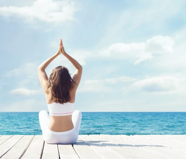 Vrouw in wit sportkleding doet yoga op een houten pier. Zee en hemel achtergrond. Yoga, sport, vakantie en reizen concept. — Stockfoto