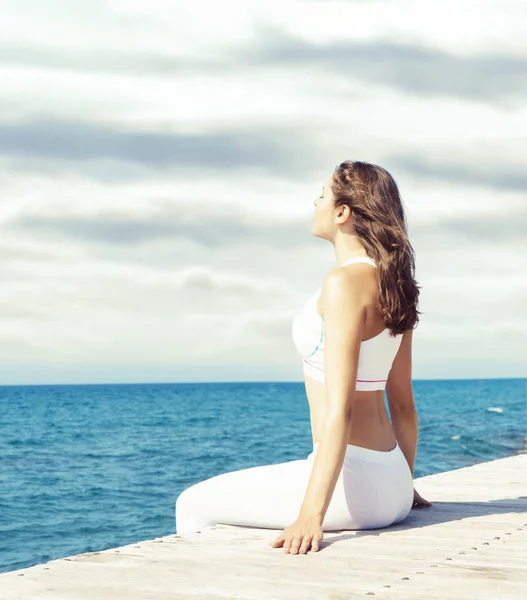 Aantrekkelijke vrouw in witte sportieve kleren doet yoga op een houten pier. Yoga, sport, ontspanning, recreatie en vrijheid concept. — Stockfoto