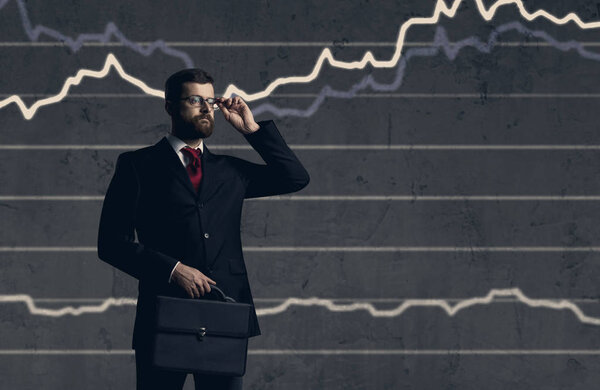 молодой и уверенный в себе бизнесмен в черном костюме стоит на тёмном фоне с бизнес-графиком и портфелем
