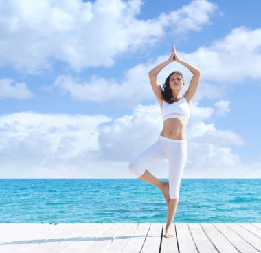 Genç ve güzel kız beyaz spor yaz ahşap bir iskele üzerinde yoga yapıyor. Yoga, spor, eğlence, tatil ve seyahat kavramı.