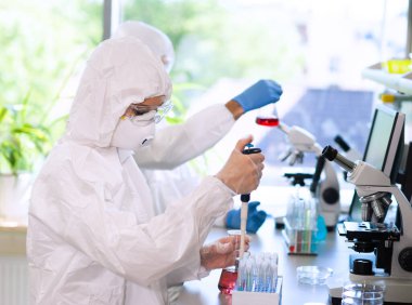 Scientists in protection suits and masks working in research lab using laboratory equipment: microscopes, test tubes. Medicine, infection and vaccine discovery concept. clipart