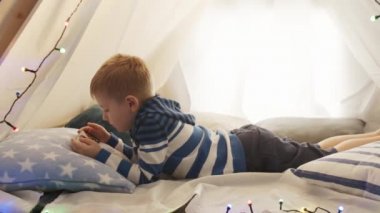 Küçük çocuk akşamları evdeki çocuk çadırında akıllı telefonuyla oynuyor. Oyun odasındaki mutlu beyaz çocuk..
