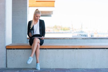 Genç ve çekici bir iş kadını ya da öğrencinin açık hava fotoğrafı. Tablet ve kahveyle gündelik giyinmiş bir kadın. İş ve eğitim kavramı.