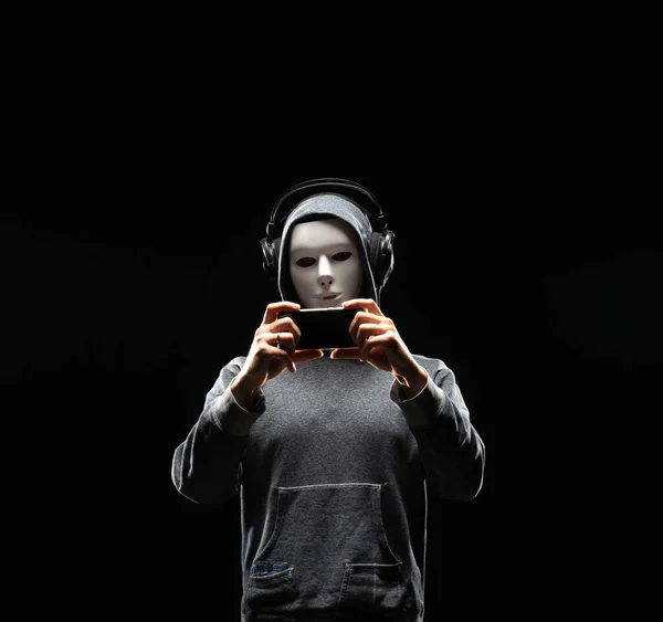 Portrait of computer hacker in white mask and hoodie. Obscured dark face. Data thief, internet fraud, darknet and cyber security concept.