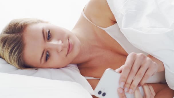 若い女性がスマートフォンでベッドの中に横たわっていた 美しい金髪の目覚めの女の子 寝室で朝 窓から日光 健康と休息の概念 — ストック動画