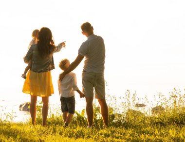 Mutlu bir aile denize yakın yürüyor. Kırsal kesimdeki tarla ve ağaçlar. Gün batımı veya gün doğumu sıcak renkler. Sevgi dolu ebeveynler ve güzel çocuklar. Aşk ve ebeveynlik kavramı.