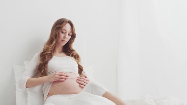 ベッドに座っている彼女の美しい腹に触れる幸せな妊娠中の女性 期待の概念 — ストック動画