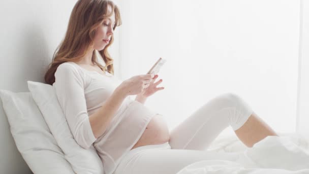 Ευτυχισμένη Έγκυος Γυναίκα Που Χρησιμοποιεί Συσκευή Smartphone Κάθεται Στο Κρεβάτι — Αρχείο Βίντεο