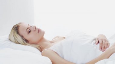 Genç bir kadın akıllı telefonuyla yatakta yatıyor. Güzel sarışın uyanış kızı. Yatak odasında sabah, pencereden gün ışığı. Sağlık ve dinlenme kavramı.