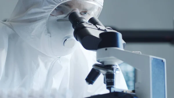 Scientist in protection suit and mask working in research lab using laboratory equipment: microscopes, test tubes. Coronavirus 2019-ncov hazard, pharmaceutical discovery, bacteriology and virology