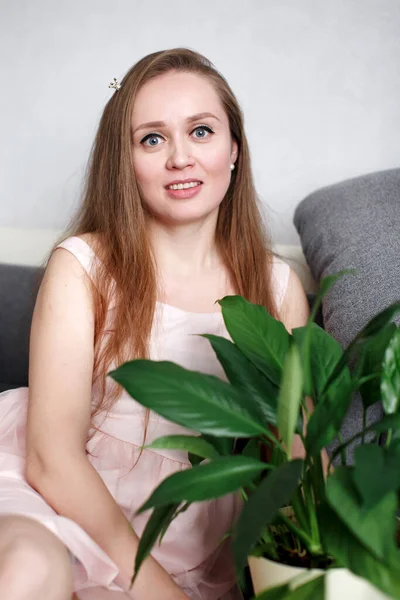 Портрет молодої щасливої жінки вдома, сидячи на дивані з зеленою свіжою кімнатною рослиною, посміхаючись — стокове фото