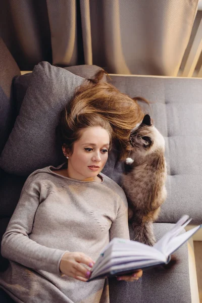 머리가 긴 매력적 인 젊은 여자가 흥미 로운 책을 읽으면서 집에서 고양이와 함께 쉬고 있습니다 — 스톡 사진