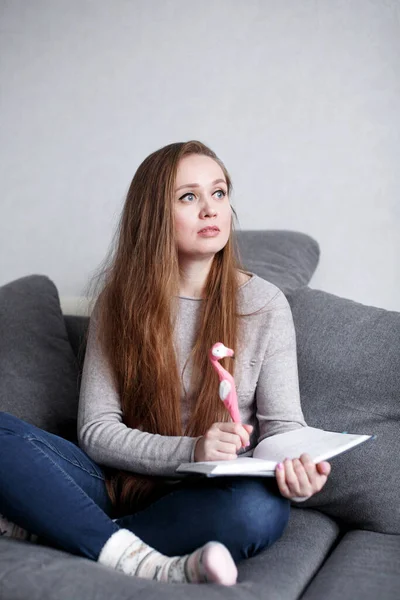 Творческая задумчивая молодая женщина сидит дома на диване и пишет что-то в своем дневнике смешной розовой ручкой в виде фламинго. Привлекательная женщина с длинными волосами . — стоковое фото