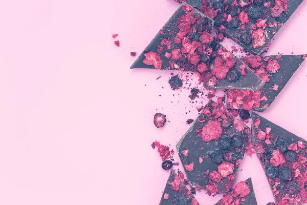 Gebrochene schwarze Schokoriegel mit verstreuten getrockneten roten Beeren auf rosa Hintergrund, flache Lage mit Platz — Stockfoto