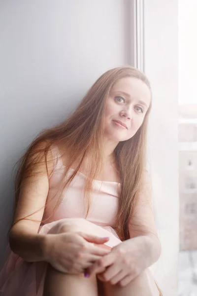 Mujer atractiva joven con hermoso pelo largo sentado cerca de la ventana mirando a la cámara sonriente, mujer real, cara natural — Foto de Stock