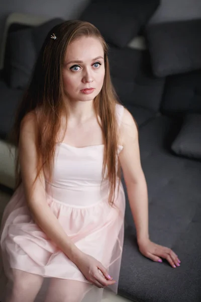 Портрет природної красивої вдумливої жінки з сірими очима і довгим волоссям, озираючись, одягнений в рожеве плаття, сидить на дивані — стокове фото
