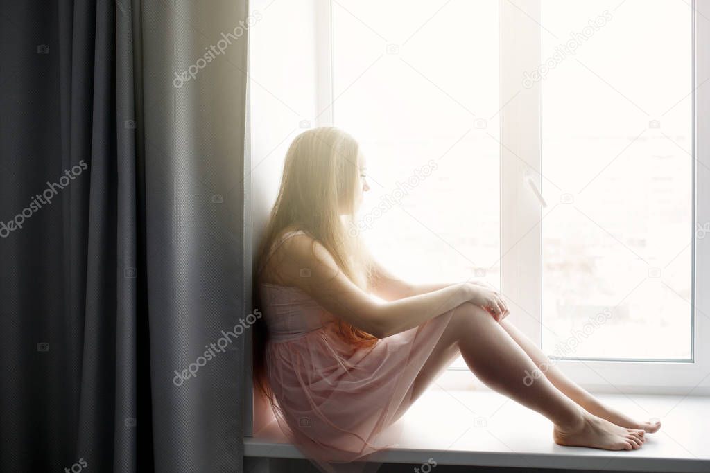 Young woman with long hair sitting at the window indoors barefoot, looking out the window, full lenght
