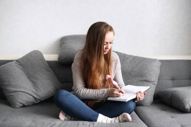 Yaratıcı, düşünceli genç bir kadın. Evdeki kanepede oturup günlüğüne bir şeyler yazıyor. Tam vücut portresi. Mesafe öğrenme veya çalışma kavramı.