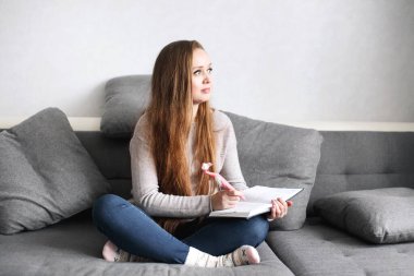 Yaratıcı, düşünceli genç bir kadın. Evdeki kanepede oturup günlüğüne bir şeyler yazıyor. Tam vücut portresi. Mesafe öğrenme veya çalışma kavramı.