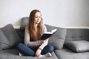 Evdeki kanepede oturup kitap okuyan zeki genç bir bayan. Modern minimalist iç mekanda uzun saçlı çekici bir kadın. Tam vücut portresi. Mesafe öğrenme veya çalışma kavramı.