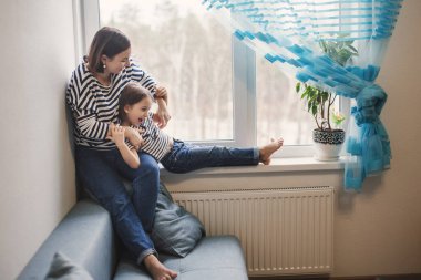 Mutlu genç bir anne, küçük kızıyla pencerenin yanında eğleniyor. Kısa esmer saçlar, aynı kıyafetler, gülümseyen ve kızını gıdıklayan bir kadın.