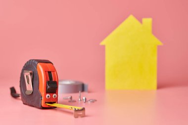 House renovation funny concept. Metal tape measure and other repair items. Home repair and redecorated concept. Yellow house shaped figure on pink background.