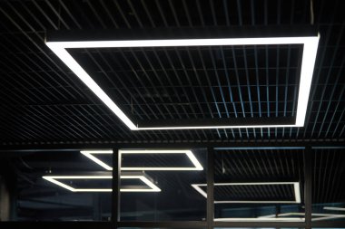 Modern ofis ışıklandırması. Ofisin karanlık tavanında ince lambalar. Beyaz soğuk ışığı işyerlerinin üzerine sürdü..