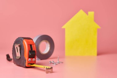 House renovation funny concept. Metal tape measure and other repair items. Home repair and redecorated concept. Yellow house shaped figure on pink background.