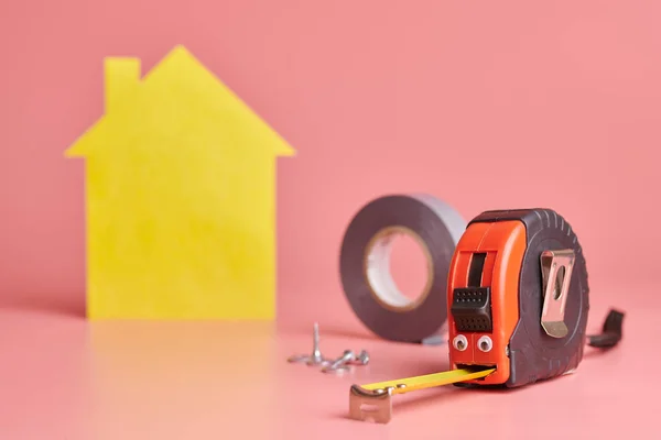 Metal Tape intézkedés vicces fogalom. Ház felújítása. Home javítási és felújított koncepció. Sárga ház alakú alak rózsaszín háttér. — Stock Fotó