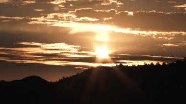 Sun becomes bright as it rises above forest hillside and warm lit clouds.
