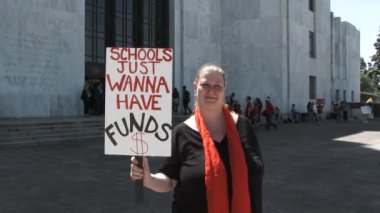 Teacher standing outside the State Capitol building holds sign reading Schools Just Wanna Have Funds.