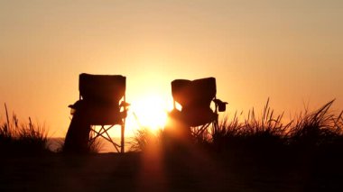 Gün batımında okyanustaki kum tepelerinde iki plaj sandalyesi boş oturur..