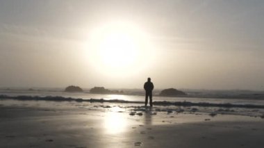 Siluetli kişi kumlu sahilde duruyor. Oregon sahilinden gün batımını seyrediyor. Sonra kameraya doğru yürüyor..