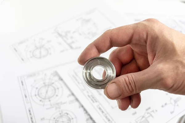 Die Hand des Ingenieurs hält ein Metallteil vor dem Hintergrund technischer Zeichnungen. Details zur Kontrollqualität. — Stockfoto
