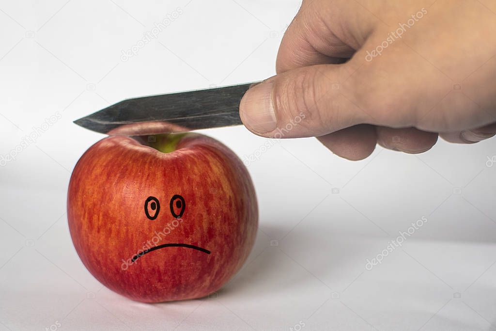 man cuts a red apple with a knife. sad face image on an apple