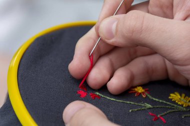 Küçük sarı bir plastik çerçevede küçük bir nakış desenli bir kız / kadın / kadının elleri. Örgü yaprağı deseni, nakış desenleri, çerçeve ve arka planda çok renkli iplikler.