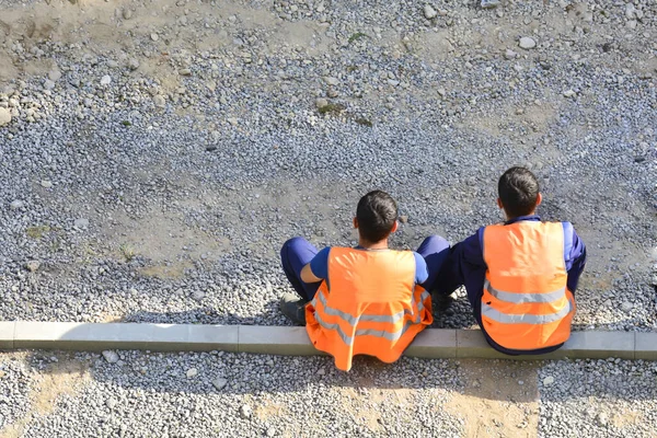 Migrants workers in yellow and orange vests resting by the road. They are sitting on the sidelines. Repair the road. Copy space. The concept of a break for rest, lack of work, unemployment.