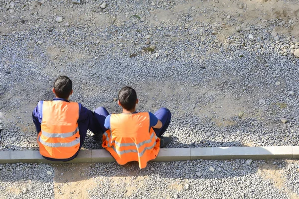 Migrants workers in yellow and orange vests resting by the road. They are sitting on the sidelines. Repair the road. Copy space. The concept of a break for rest, lack of work, unemployment.