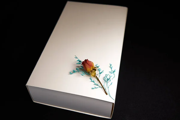 White box with a dry pink rose and a blue branch on a black background for a gift                       