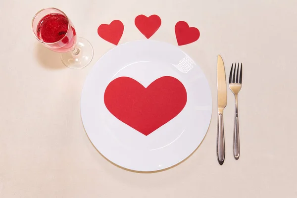 Big red heart made of paper on a white plate around a small heart with a fork and knife and a glass of champagne   for valentines day             