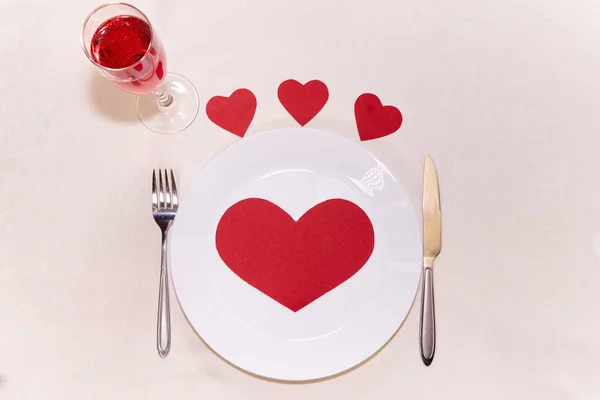 Big red heart made of paper on a white plate around a small heart with a fork and knife and a glass of champagne   for valentines day                    