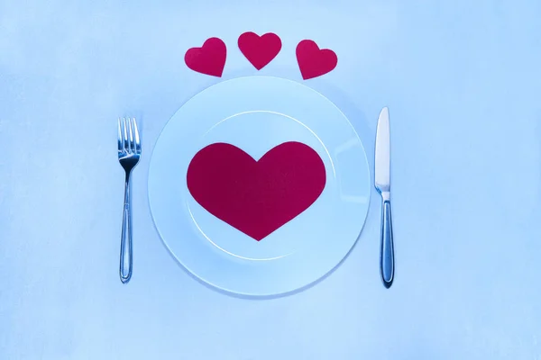Big red heart made of paper on a white plate and small hearts with a knife and a fork for Valentine's Day                             