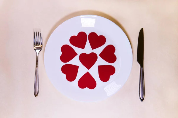  Many little red hearts on a white plate on a table with a knife and fork for valentines day                                   